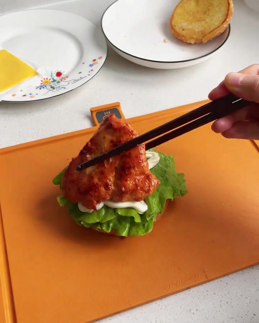 A piece of cooked, glazed chicken being placed onto a burger bun base with lettuce and mayonnaise.