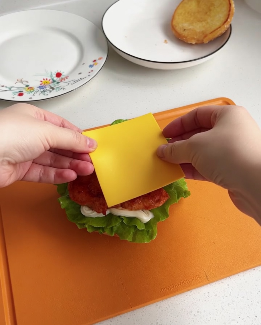 A slice of yellow cheese being centered over the hot chicken piece on the burger base.