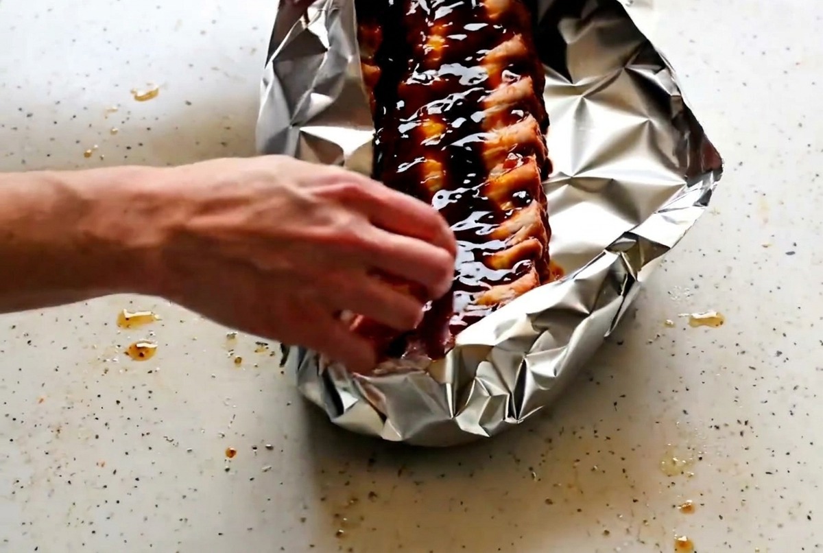 Hands carefully folding and crimping the edges of the aluminum foil to seal the pork ribs inside.