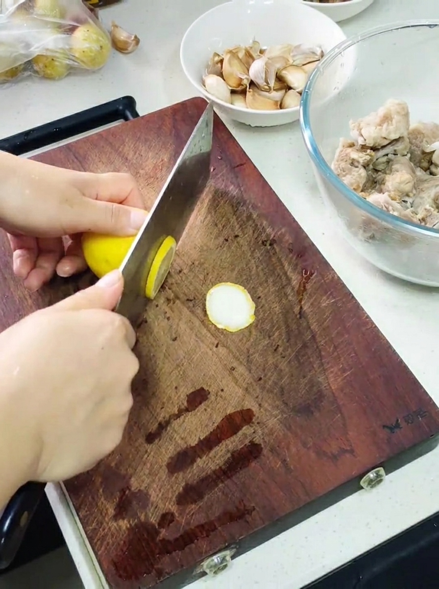 Hands using a cleaver to slice a lemon on a wooden cutting board, with a bowl of marinated ribs and garlic cloves nearby.