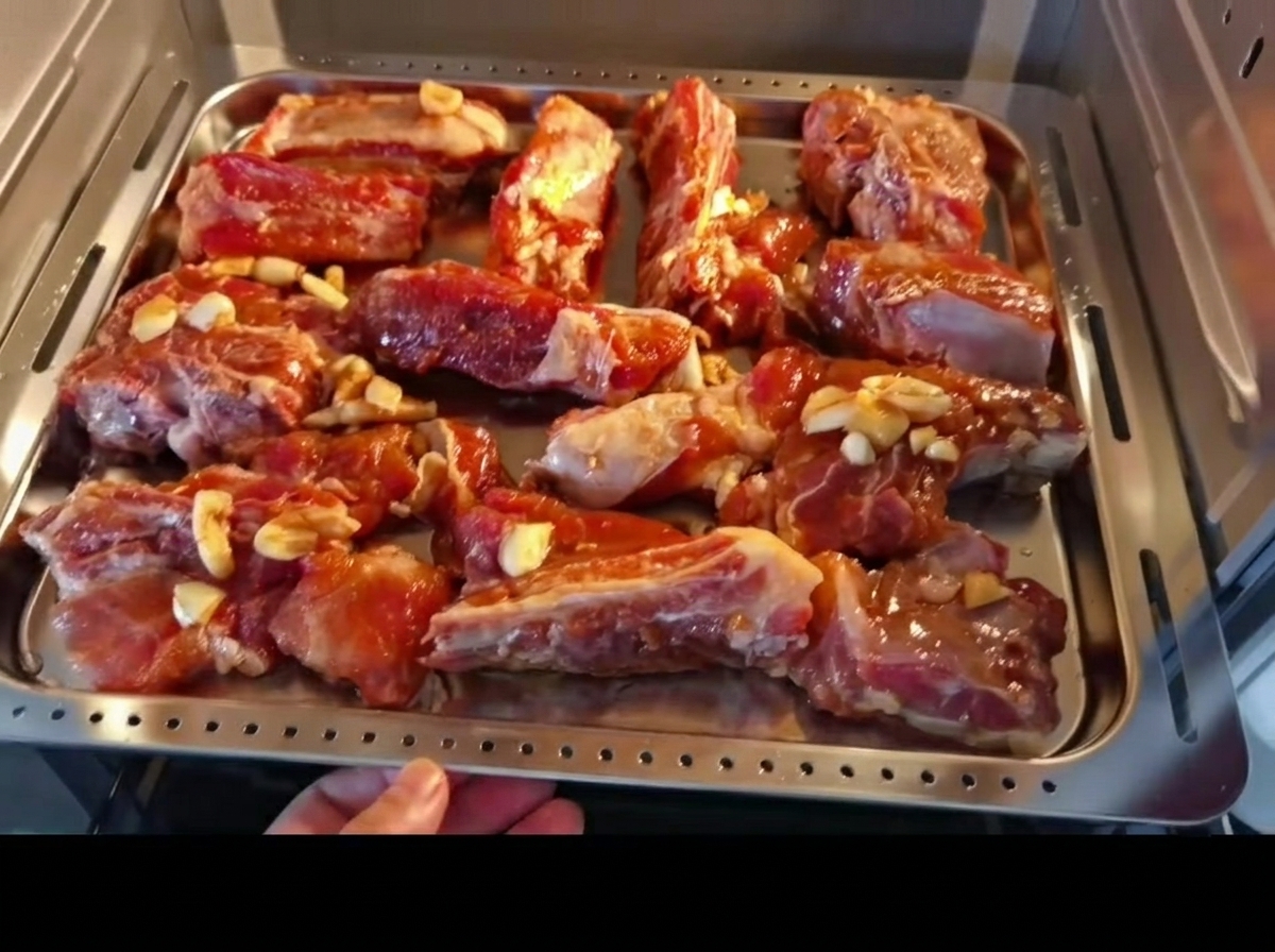 Marinated pork ribs arranged evenly on a metal baking tray, being placed into an oven.