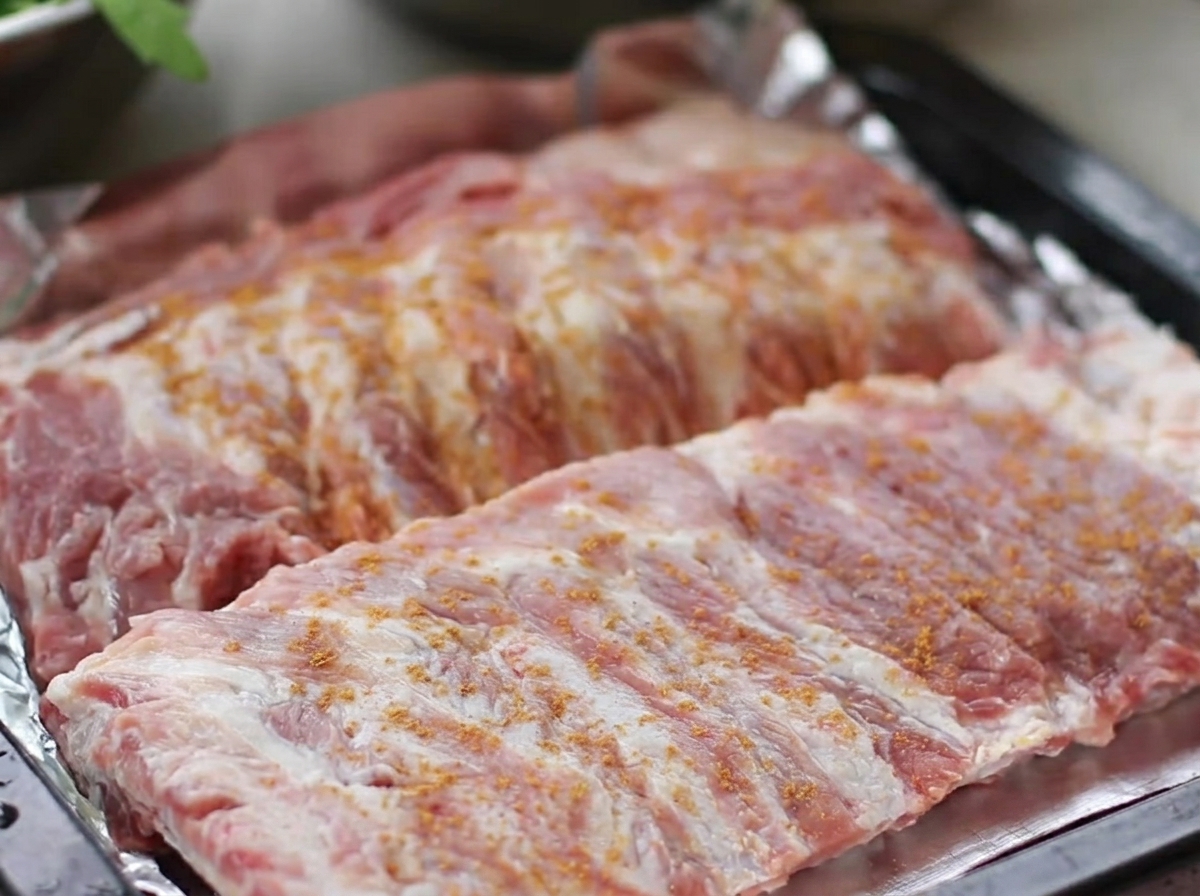 Raw pork ribs arranged in a roasting tray, generously coated with a dry rub of five-spice, black pepper, ginger, and garlic.