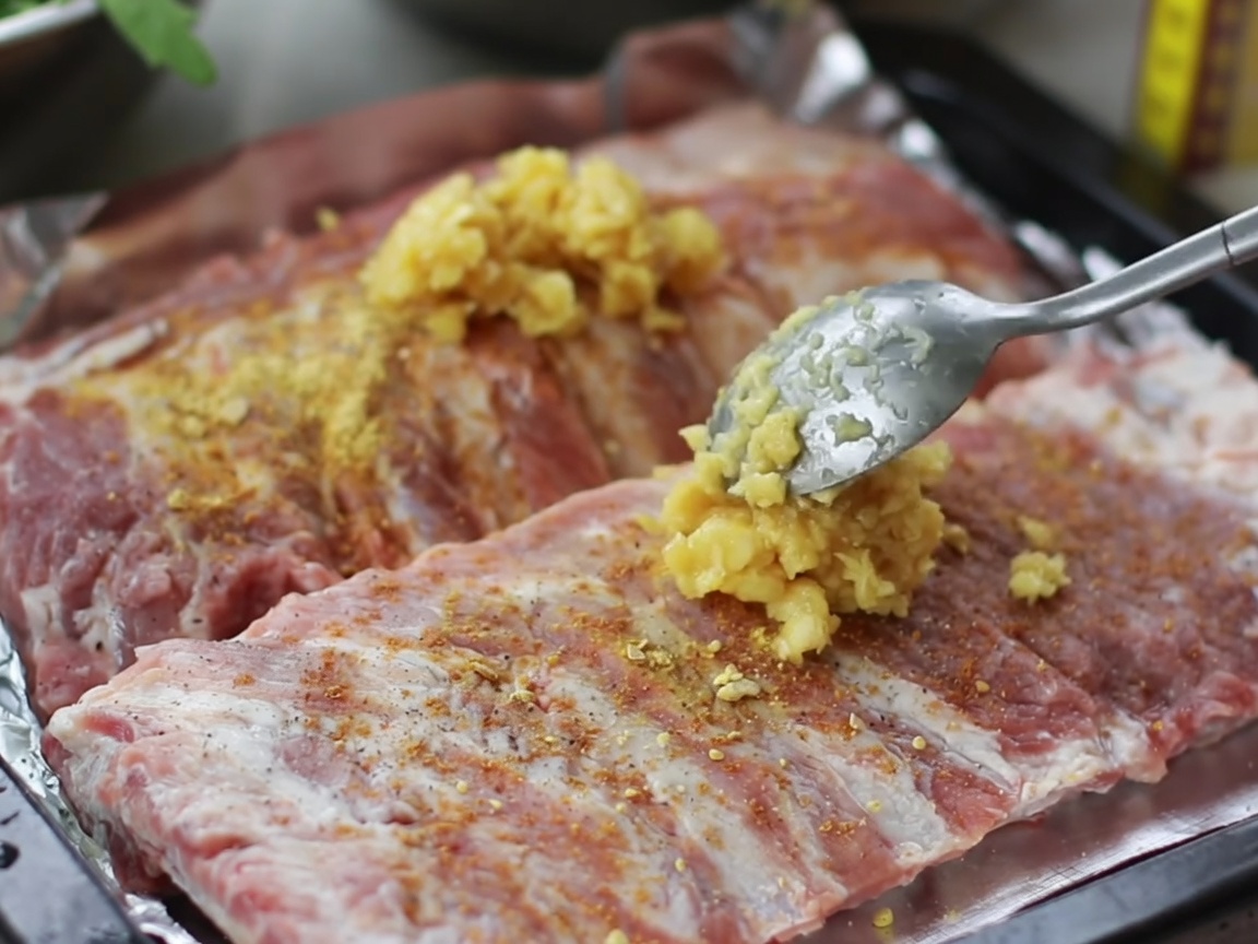 Hands rubbing a mixture of minced garlic and spices into the surface of raw pork ribs in a tray.