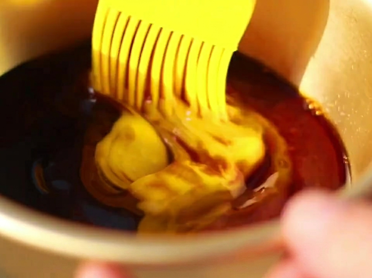A silicone brush applying a dark, rich sauce over the surface of partially roasted pork ribs in a tray.