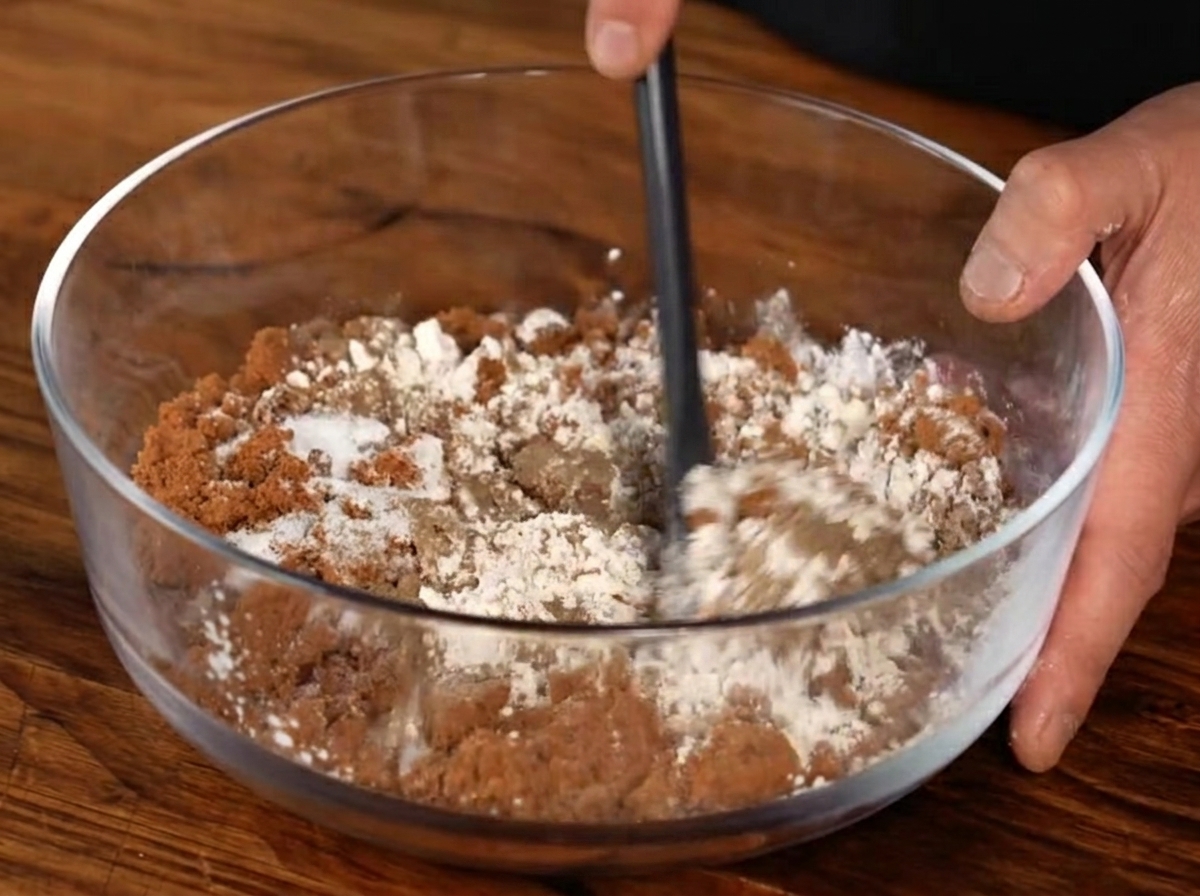 A clear bowl containing a mixture of brown sugar, garlic powder, black pepper, and salt being stirred with a black spatula.