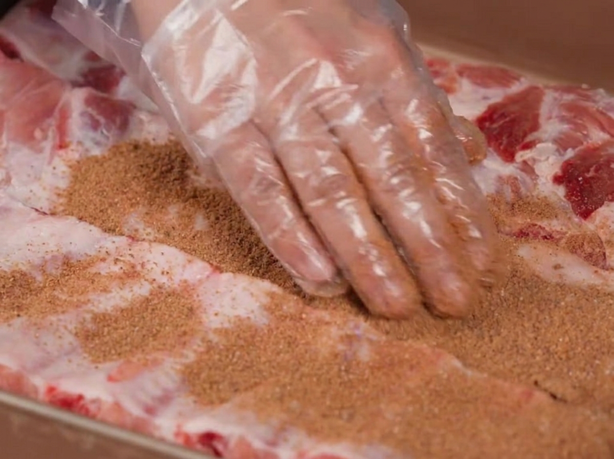 Hands wearing plastic gloves rubbing a brown spice mixture over raw pork ribs.