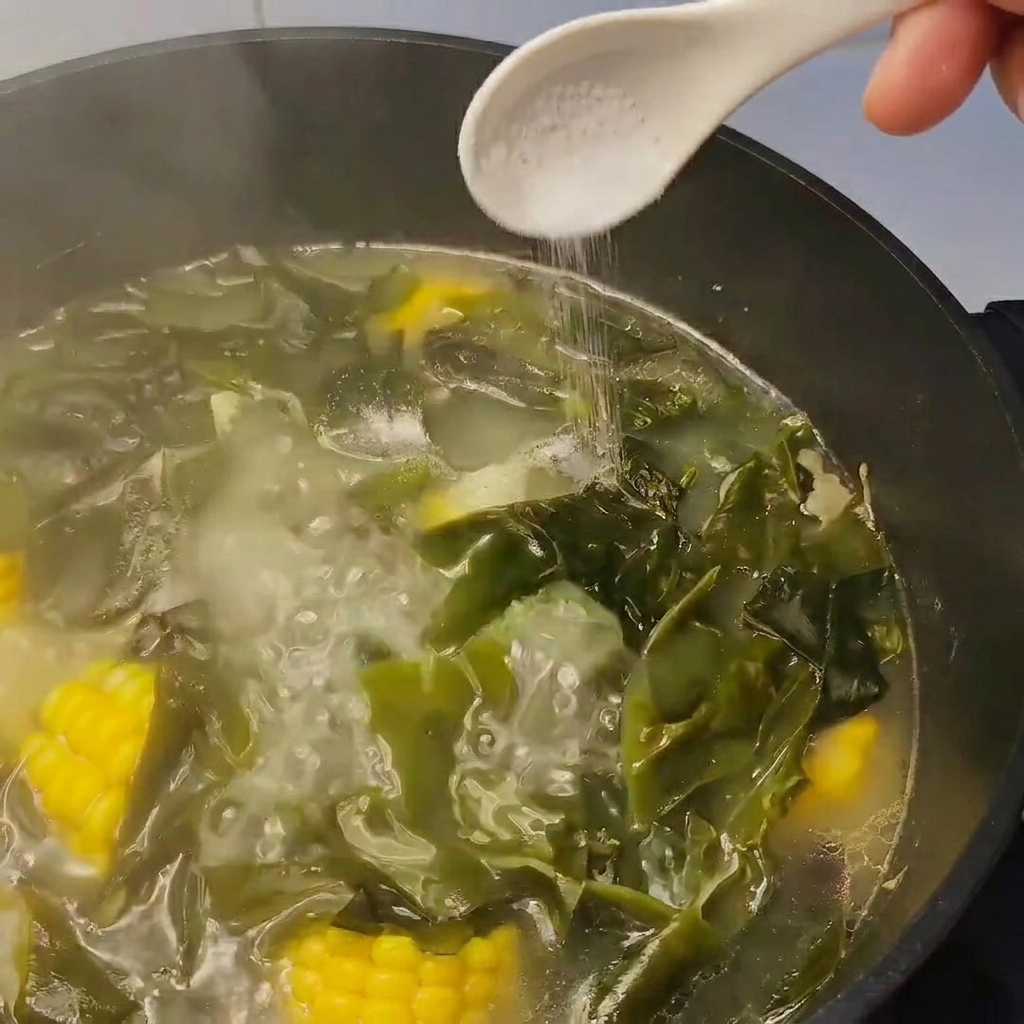 A spoonful of salt being added to a simmering pot of soup containing kelp and corn.