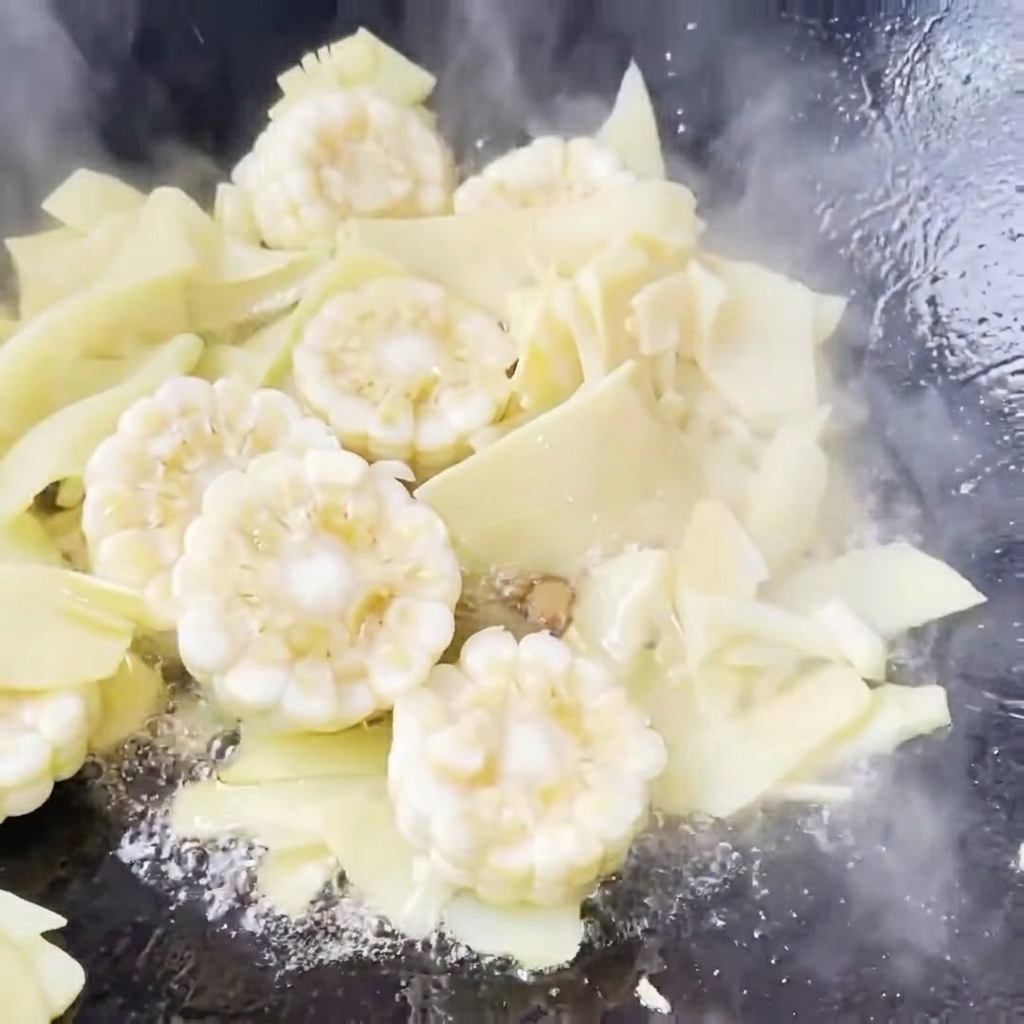 Clear water being added to a wok containing stir-fried corn and bamboo shoots, beginning to simmer.