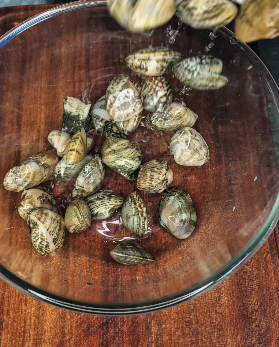 Fresh clams sitting in a clear glass bowl filled with water, salt, and a dash of oil.