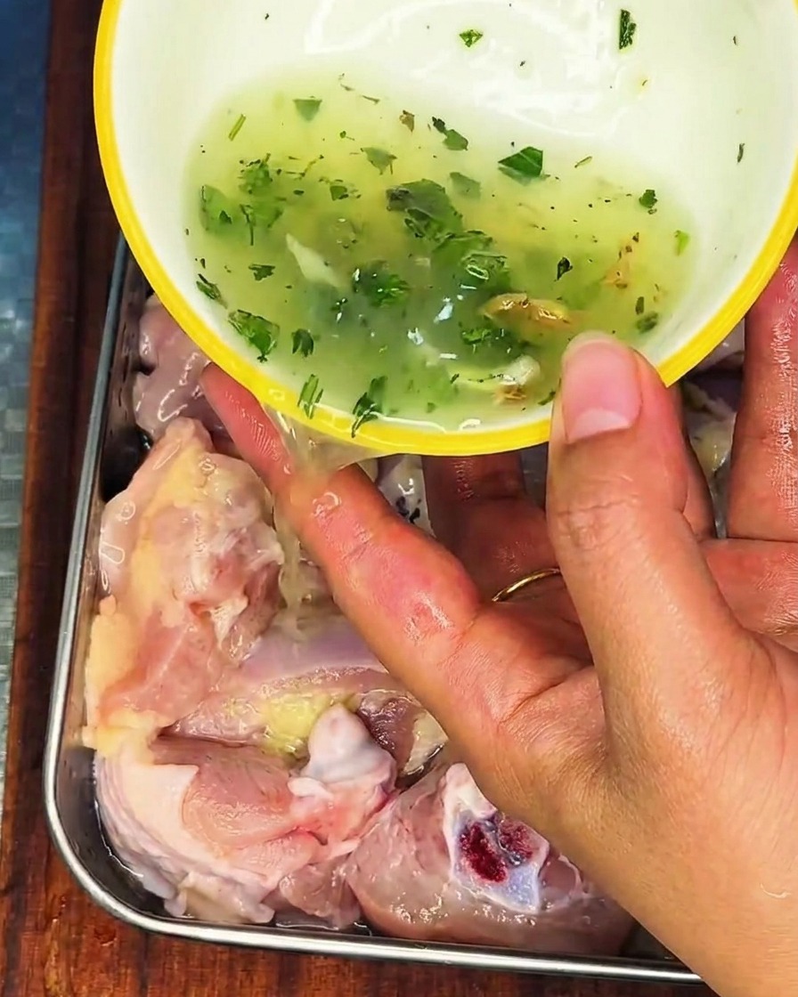 A hand pouring a green-flecked onion and ginger liquid over raw chicken in a stainless steel container.