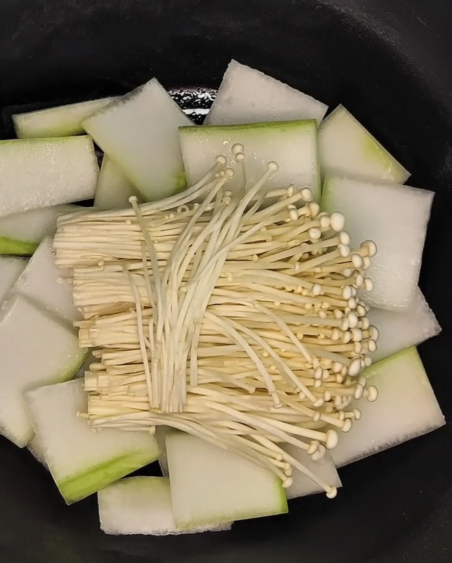 A bundle of enoki mushrooms being laid across the bed of winter melon slices in the pot.
