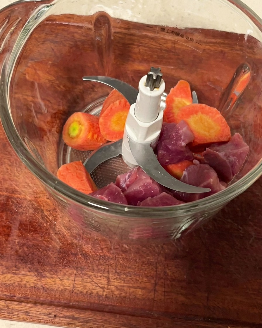 Pork pieces and carrot chunks inside a clean meat grinder container ready to be minced.