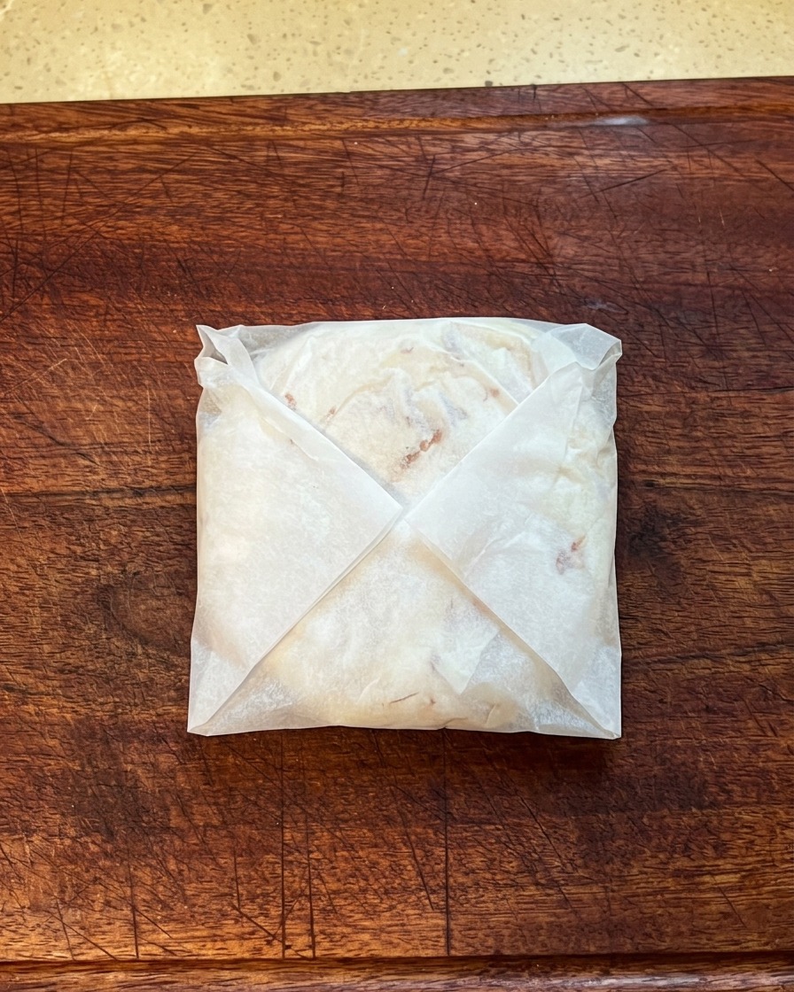 A scallion pancake folded and encased in parchment paper, being pressed flat on a wooden cutting board.