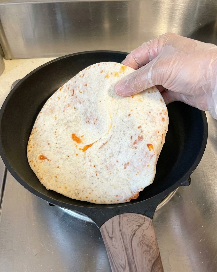 A hand wearing a plastic glove places a flattened, parchment-wrapped pancake into a hot black non-stick skillet.