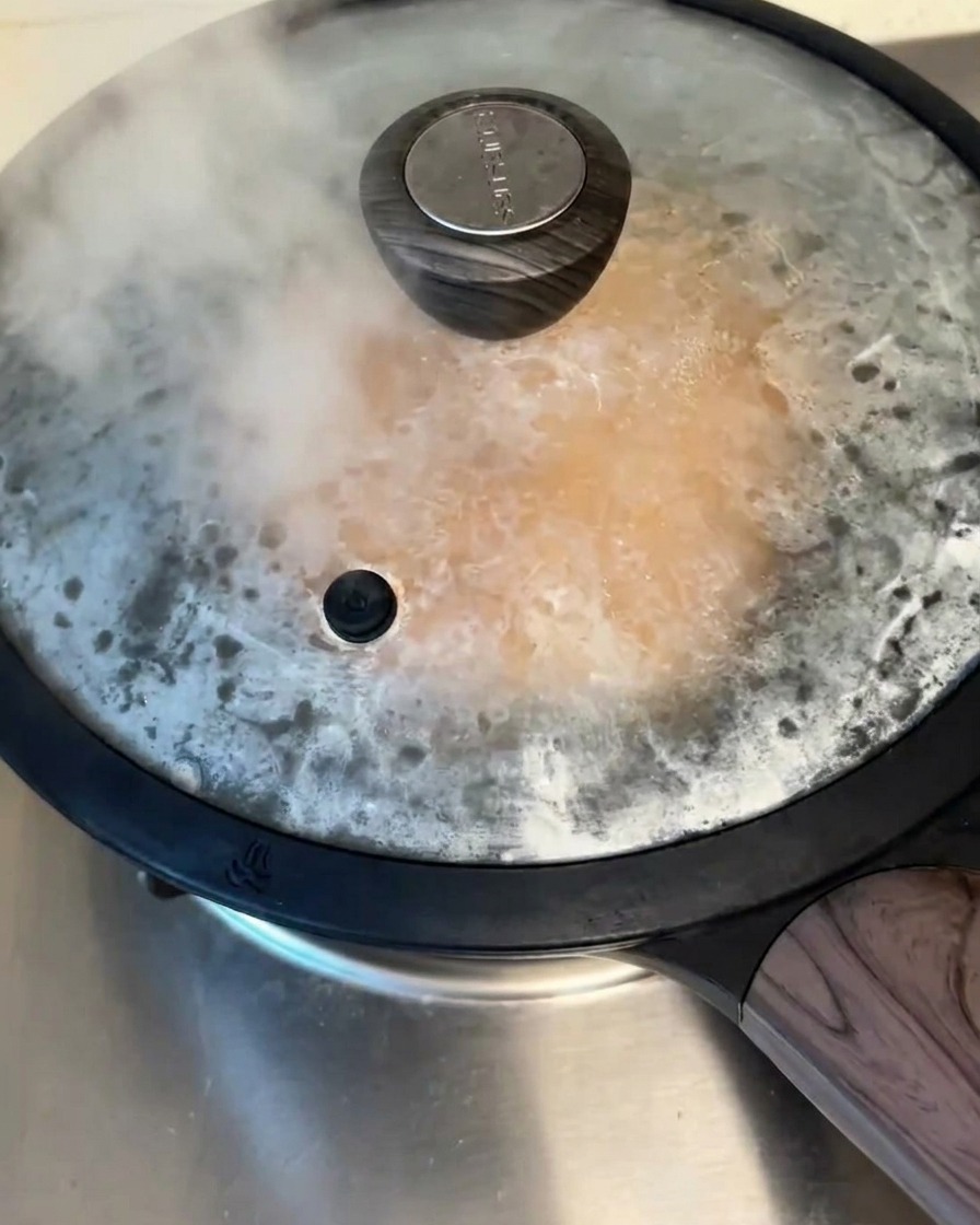 A covered non-stick pan on the stove, with visible steam escaping from under the lid while the pancake cooks.