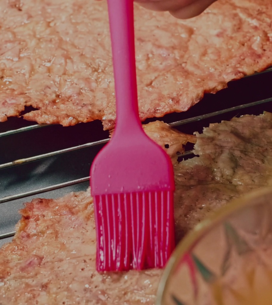 Silicone brush applying a honey-water glaze onto pork jerky slices on a wire rack.