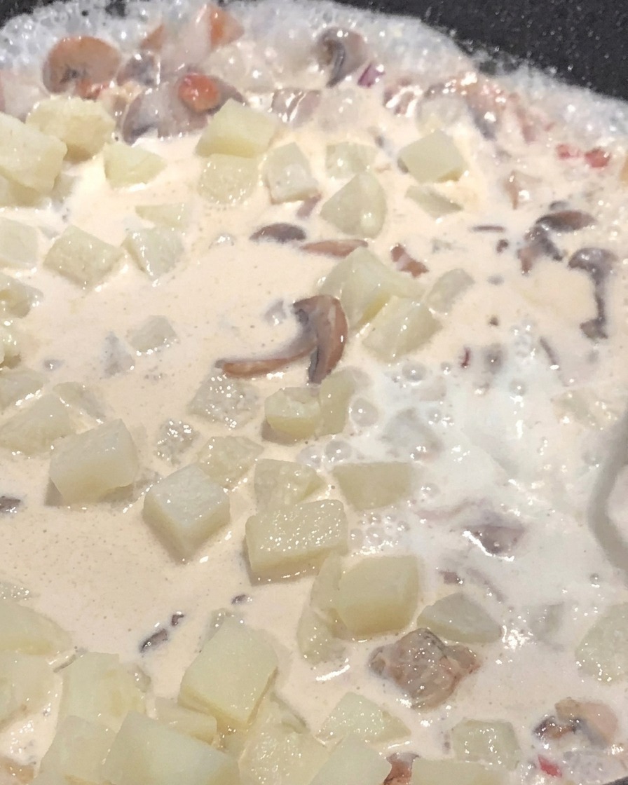 Milk being poured into a pan filled with diced potatoes and mushrooms, simmering in a creamy sauce.