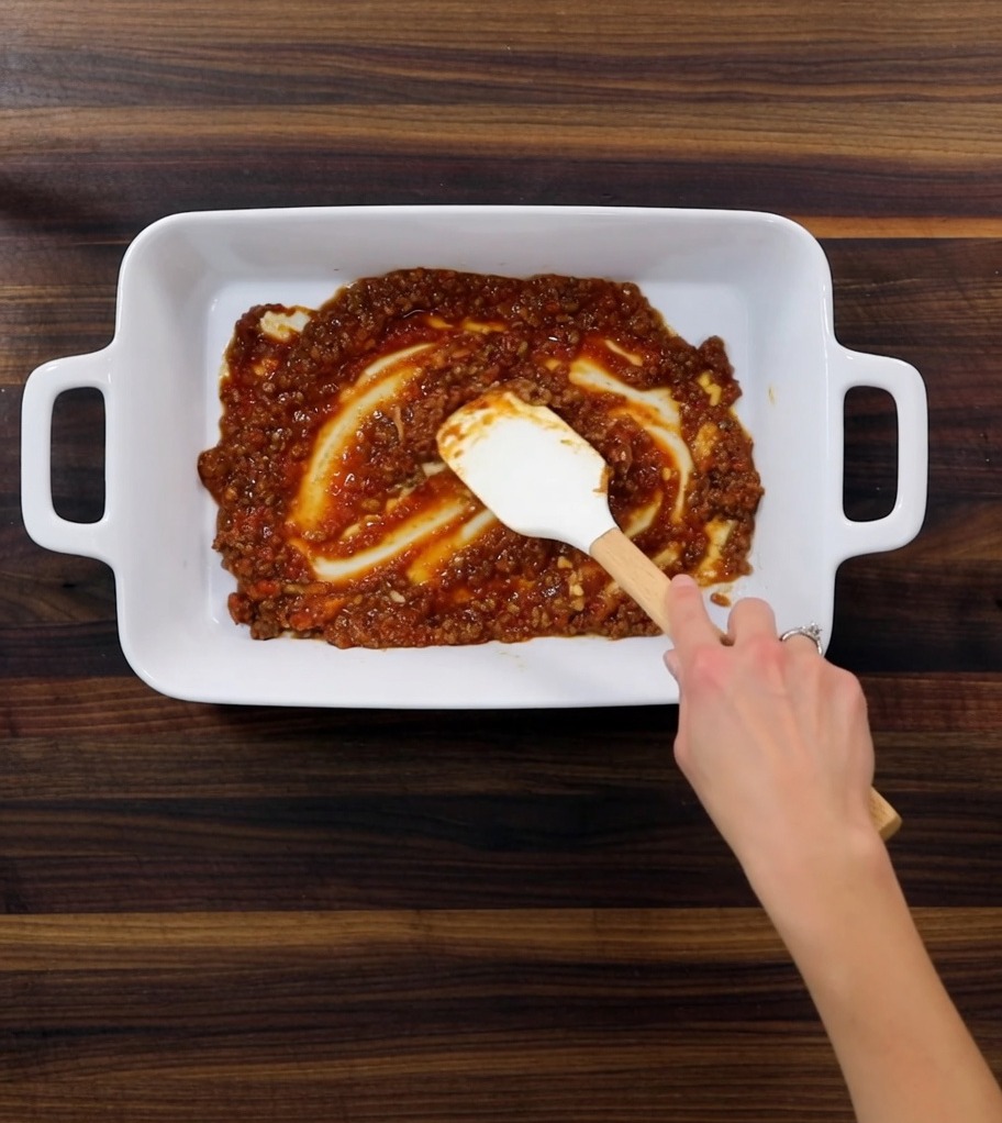 A close-up of a baking dish showing the distinct layers of savory meat sauce, tender pasta sheets, and creamy white sauce prepared for baking.
