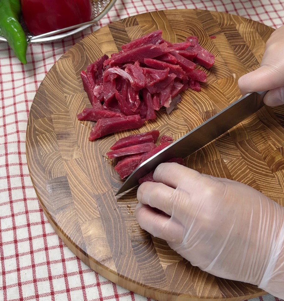 Raw beef being sliced into thin strips on a wooden cutting board.