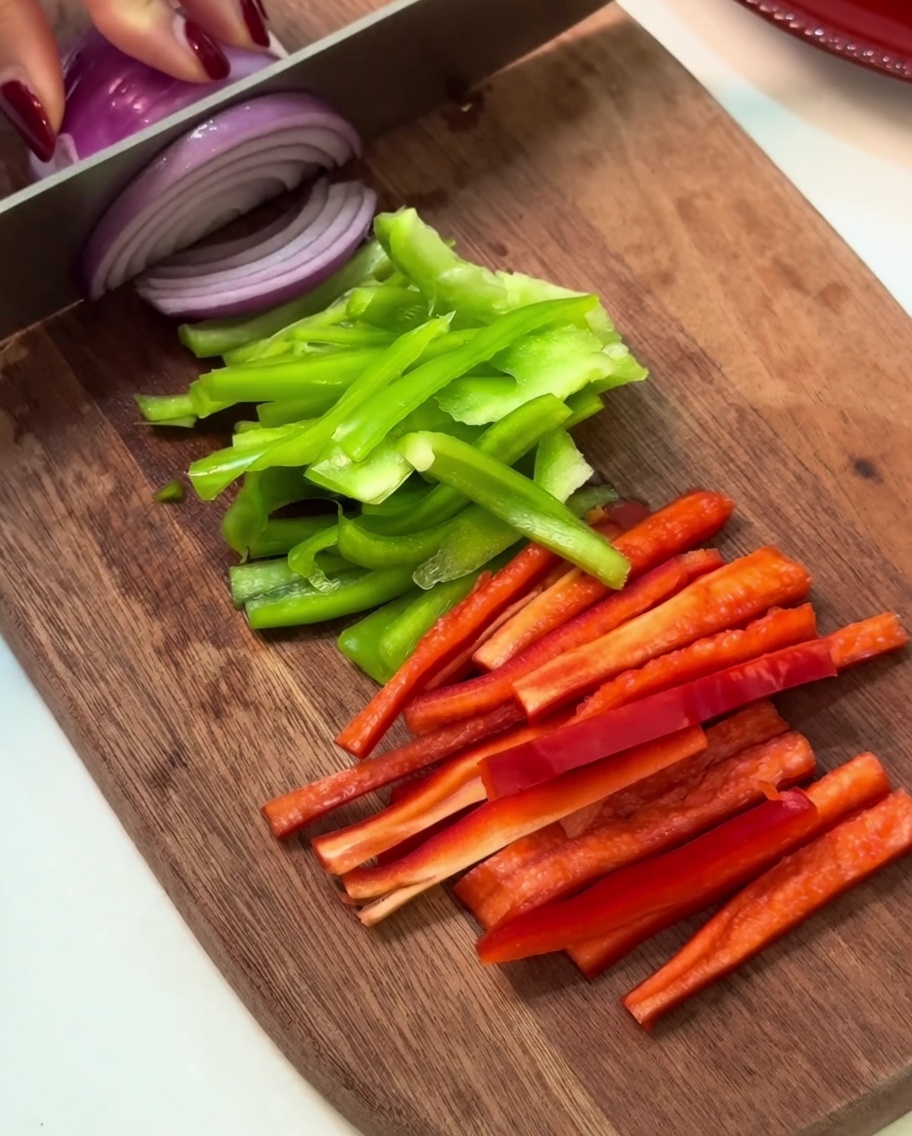 Fresh green bell pepper, red bell pepper, and purple onion slices arranged neatly on a wooden cutting board.