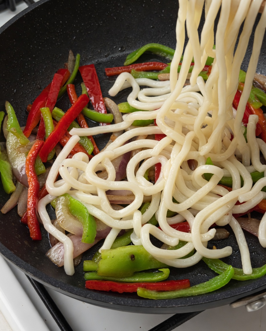 Udon noodles and beef strips being added to the pan of vegetables.