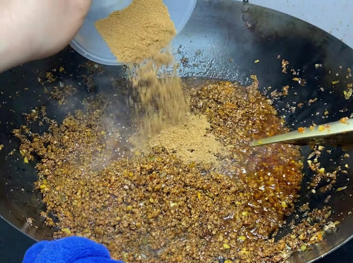 A dry spice powder mixture being sprinkled over cooked ground meat in a wok.