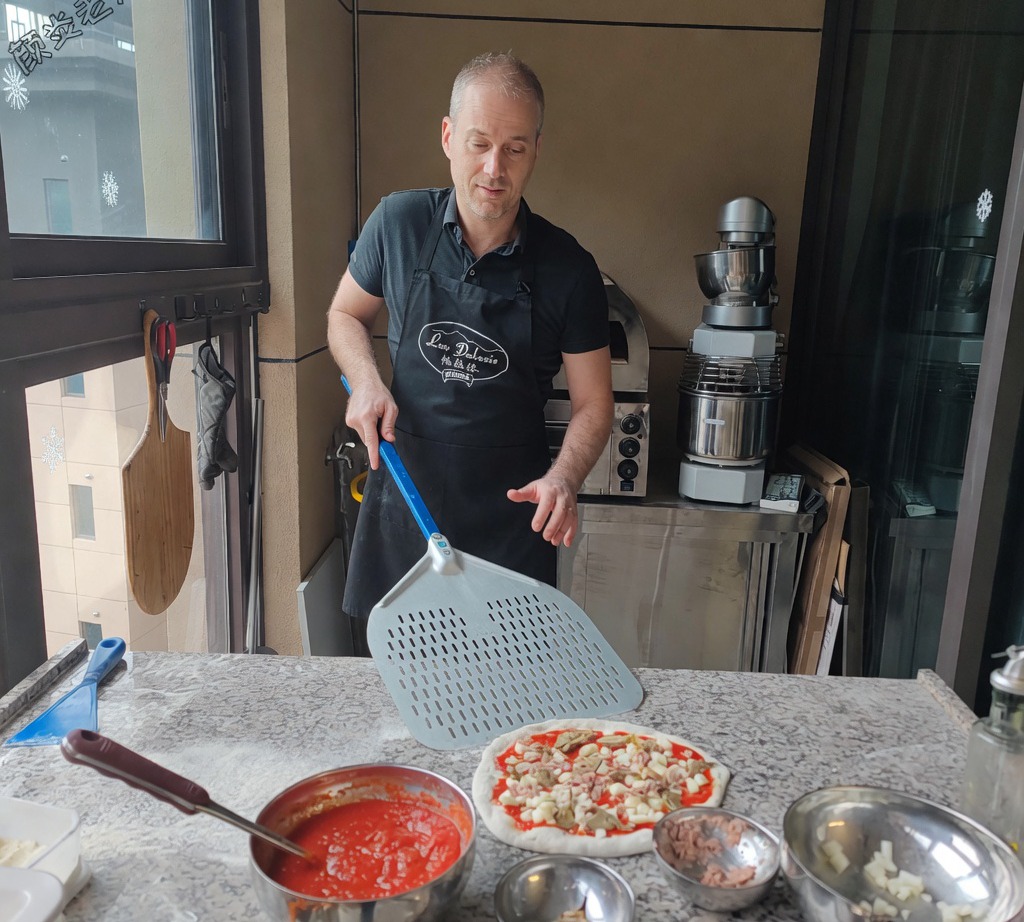 A metal pizza peel being slid under the assembled raw pizza to prepare it for baking.