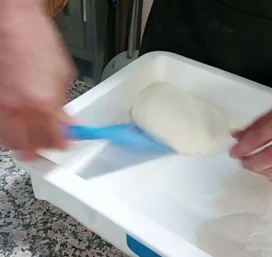 A portion of pizza dough being lifted from a container using a blue plastic scraper.