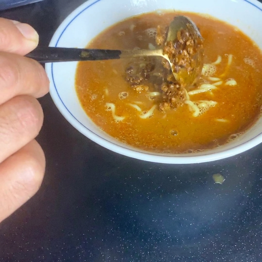A spoonful of savory brown minced meat being placed into the center of the noodle bowl.