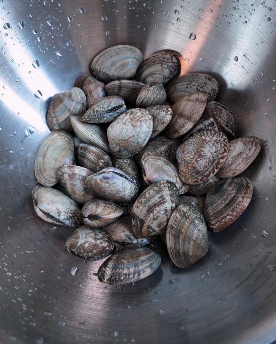 A metal bowl filled with raw, unopened clams with water droplets on the side.