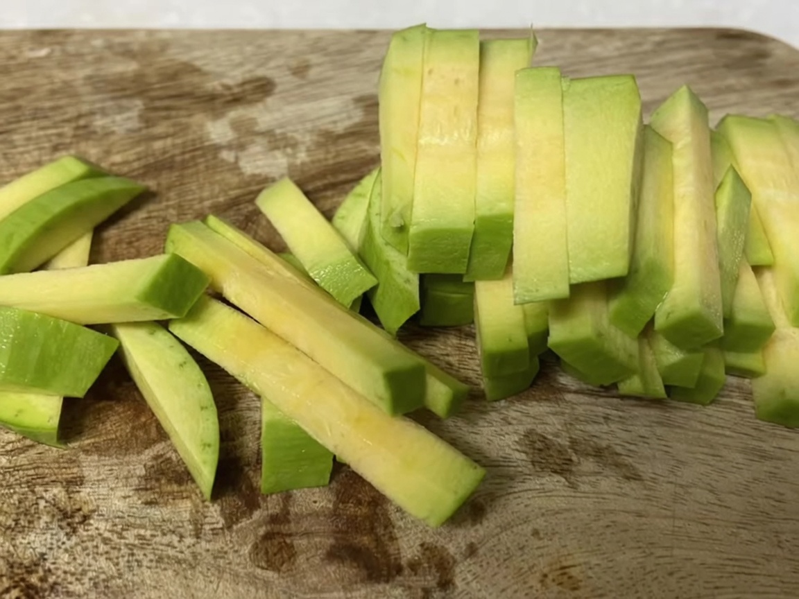 Zucchini slices stacked and being cut into thin, uniform strips on a wooden cutting board.