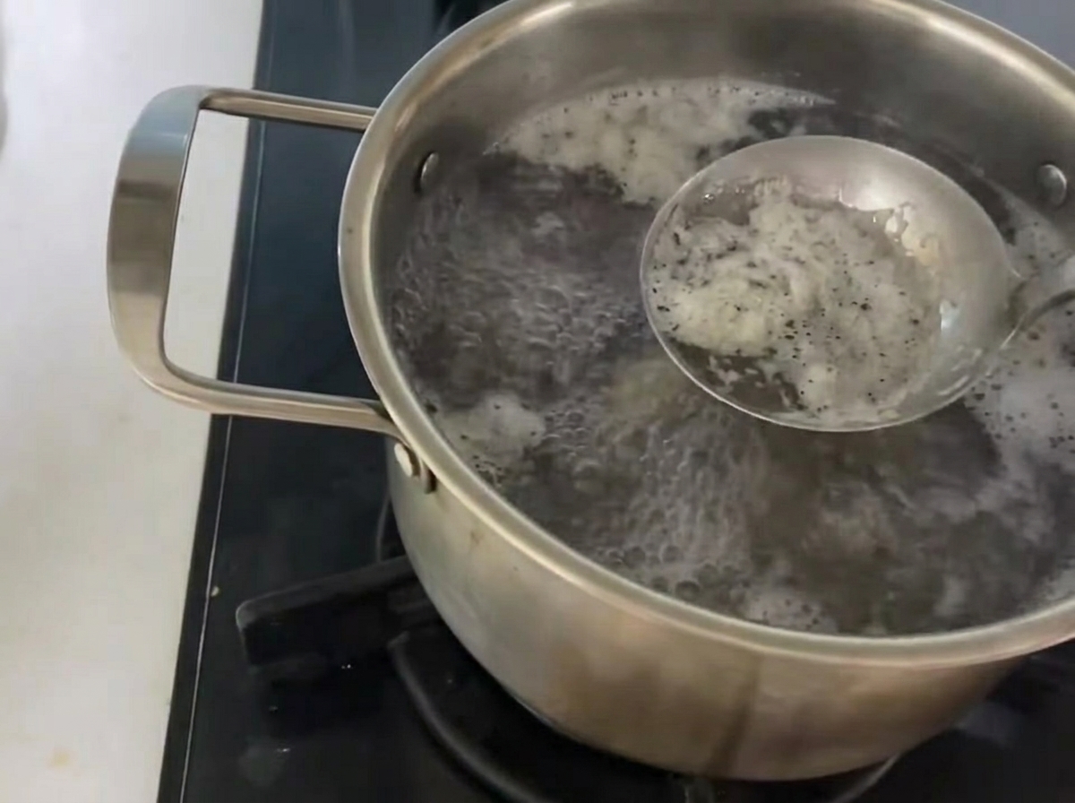A metal ladle skimming white foam from the surface of a pot of boiling broth.