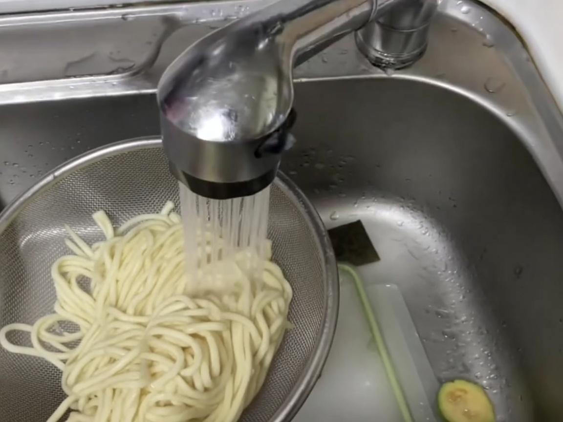 Hand-rolled noodles in a metal strainer being rinsed under a cold water tap.