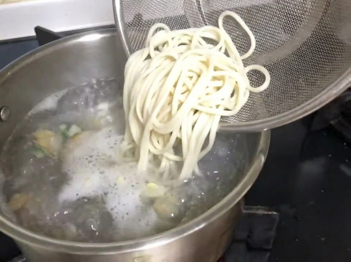 Rinsed noodles being dropped from a strainer into a pot of boiling clam broth.