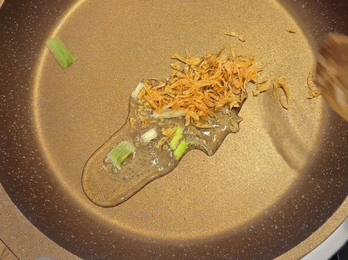 Dried baby shrimp and sliced green onions being gently stir-fried in oil in a shallow pan.