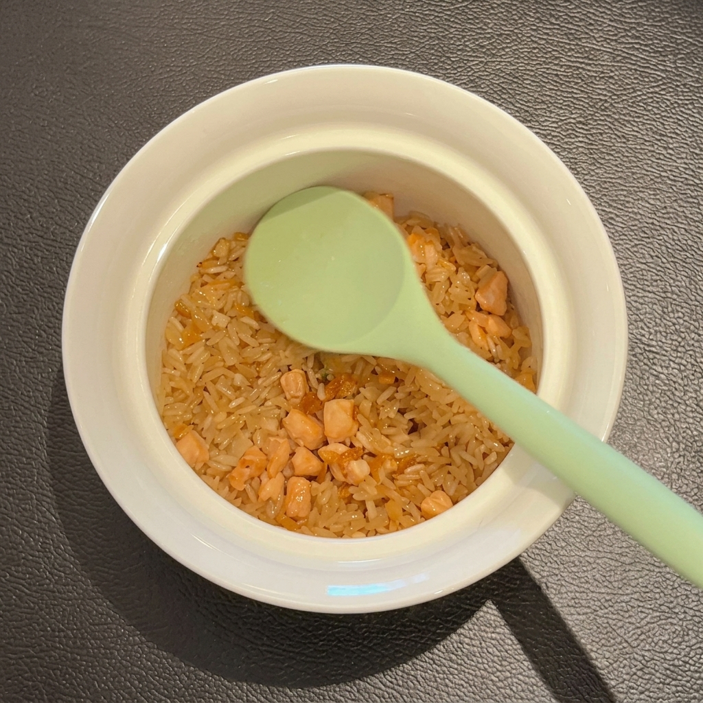 A green spoon resting inside a white ceramic bowl filled with the stir-fried rice and salmon mixture.