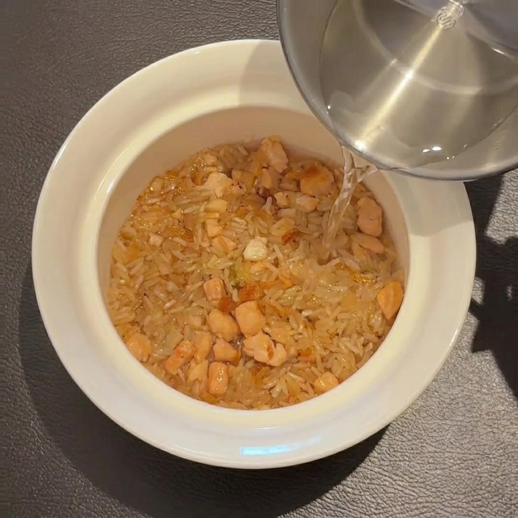 Water being poured from a stainless steel cup over the rice mixture in a white bowl.
