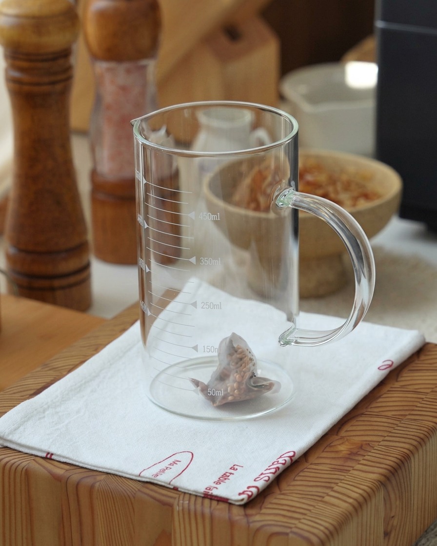 A genmaicha tea bag steeping in hot water inside a clear glass measuring cup, showing the tea beginning to infuse.