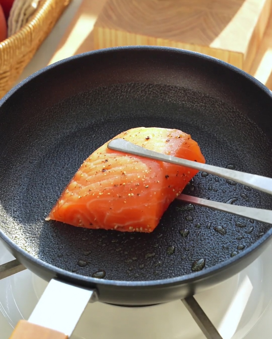 A seasoned raw salmon fillet being placed into a hot black skillet lightly coated with olive oil using metal tongs.