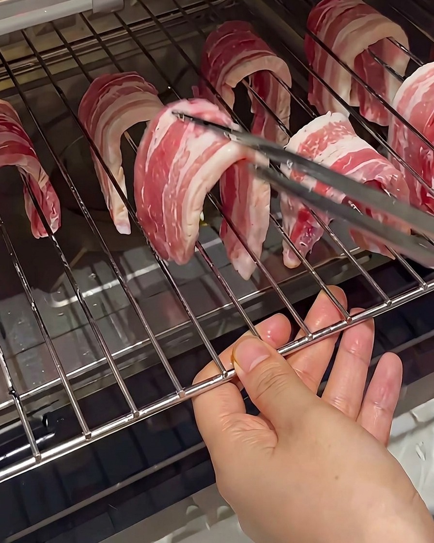 Slices of raw pork belly hanging over the metal wires of an oven rack, held by tongs.