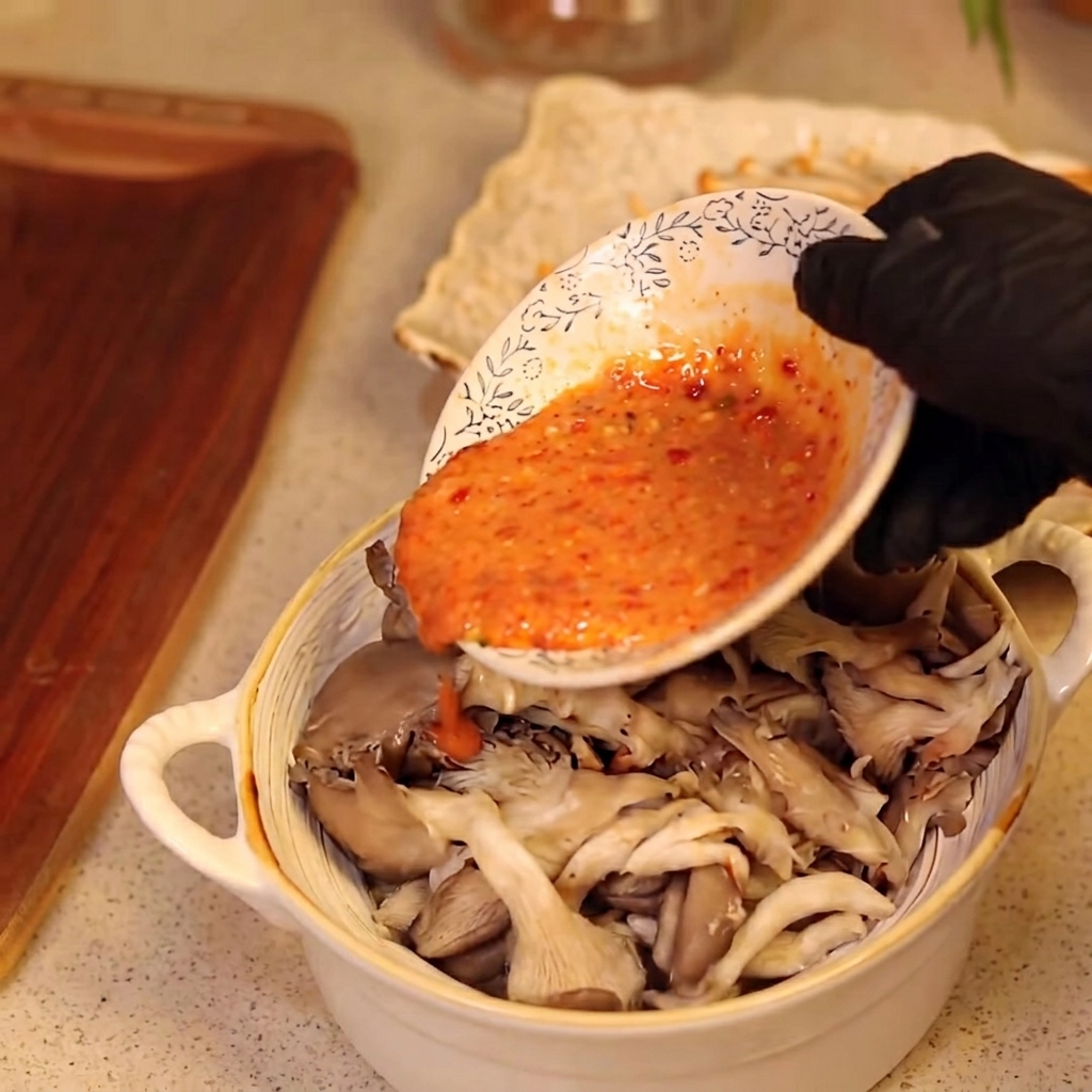 A hand wearing a black glove pouring sauce onto a bowl of mushrooms.
