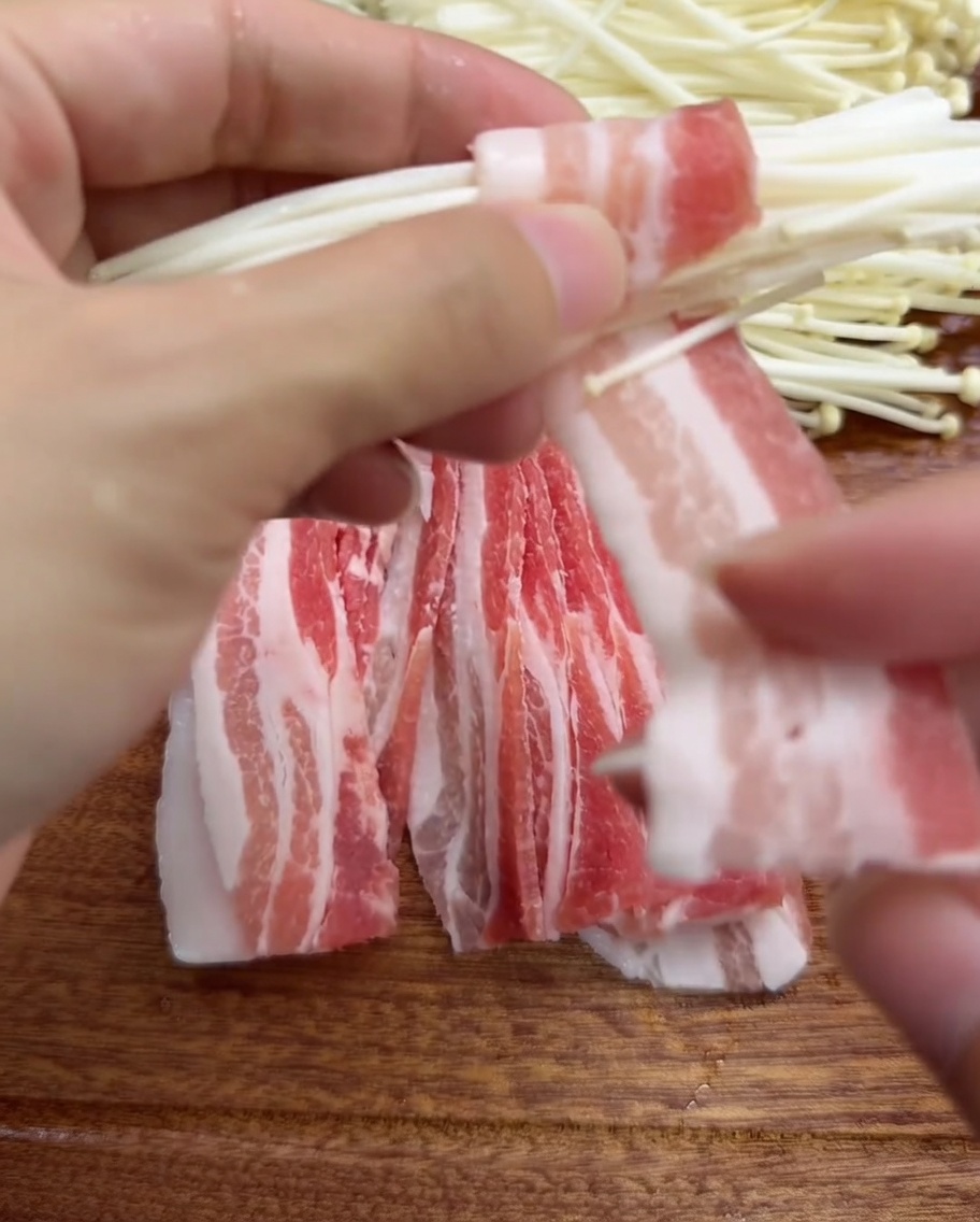 Close-up of hands wrapping a thin slice of pork belly around a cluster of enoki mushrooms.
