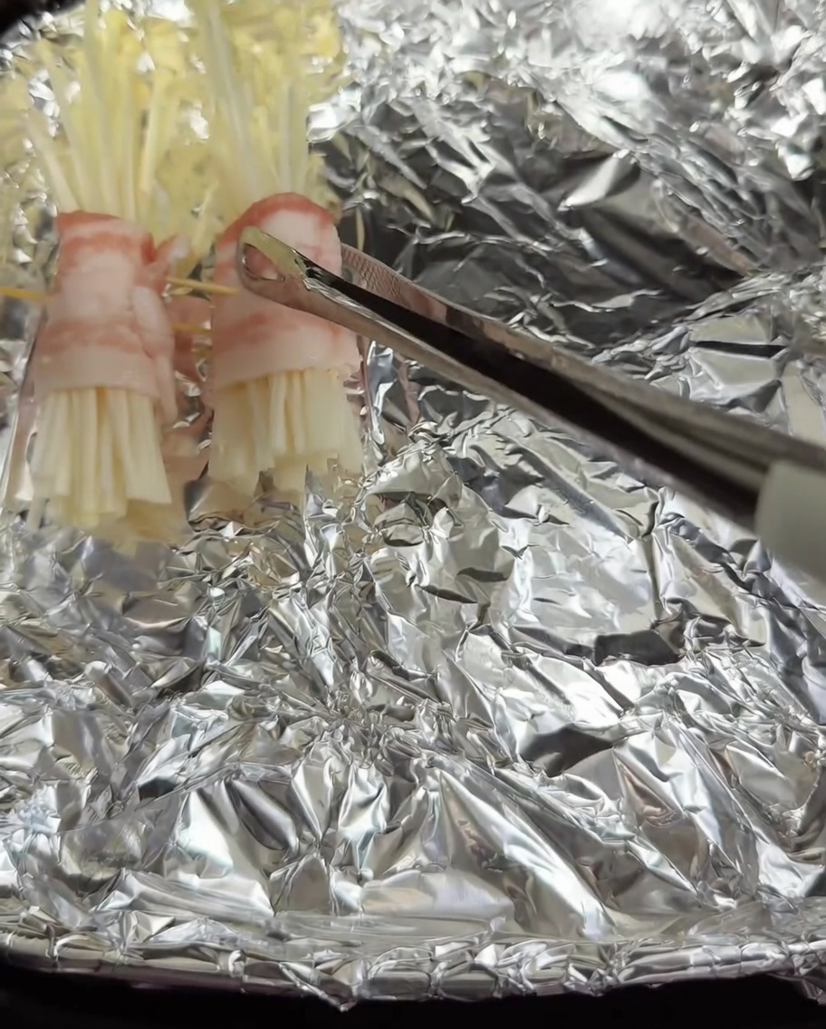 Assembled pork belly enoki rolls being placed into an air fryer basket lined with foil.