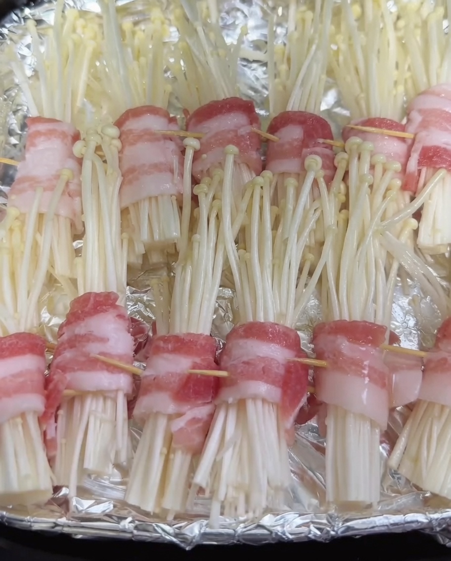 Spray bottle applying a fine mist of oil over raw pork-wrapped enoki mushroom rolls in an air fryer basket.