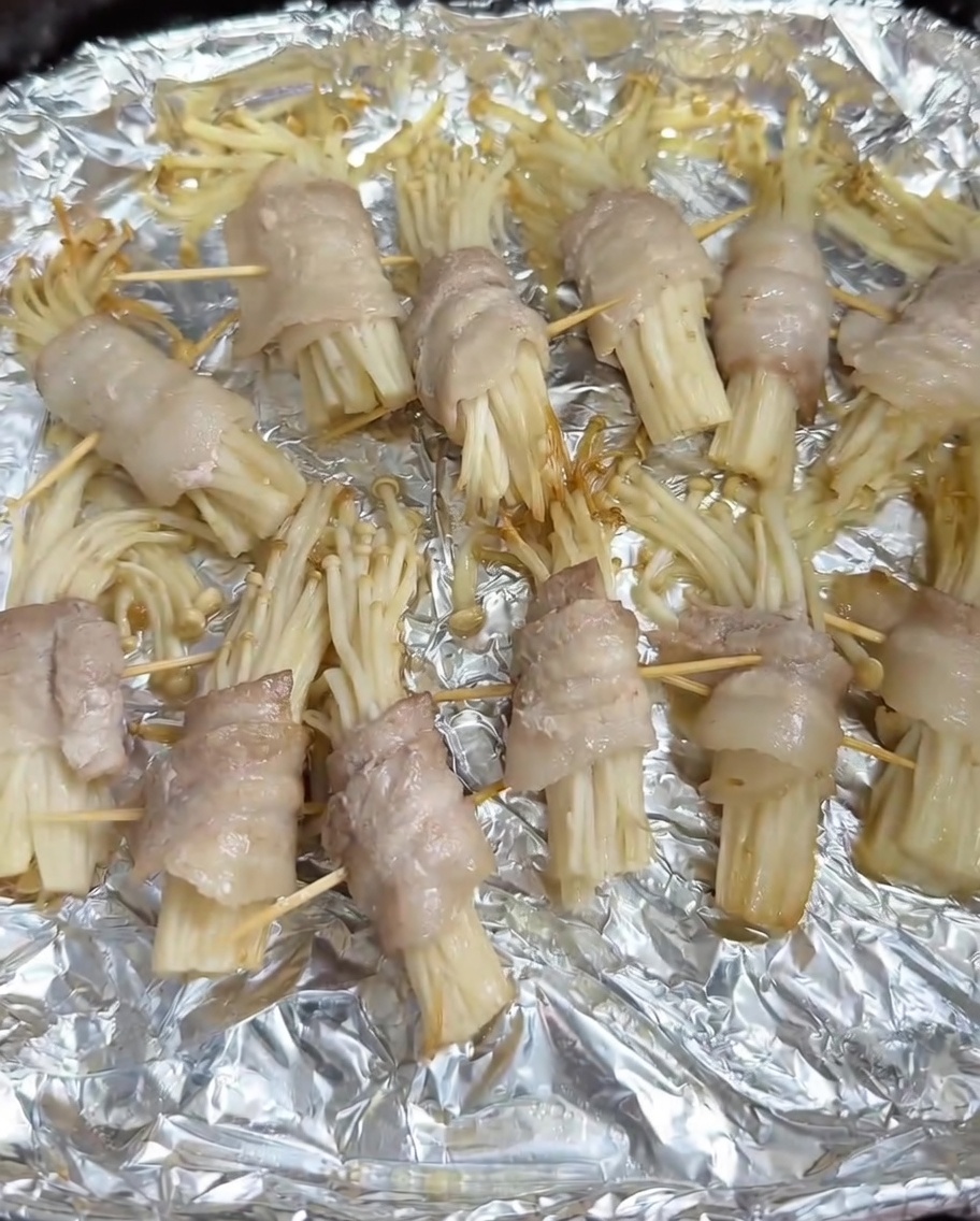 Barbecue seasoning being sprinkled onto semi-cooked pork-wrapped enoki rolls in an air fryer basket.