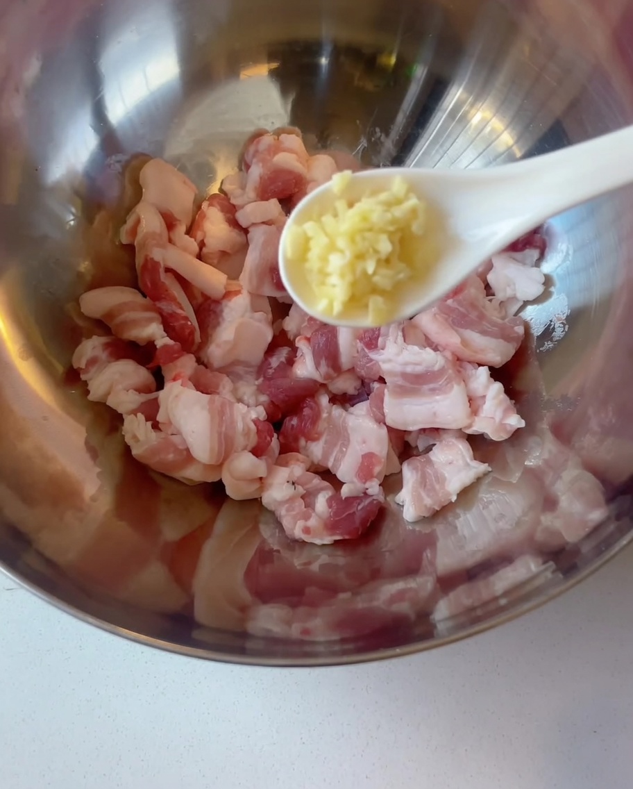 Raw pork belly slices in a metal bowl with minced garlic being added from a spoon.