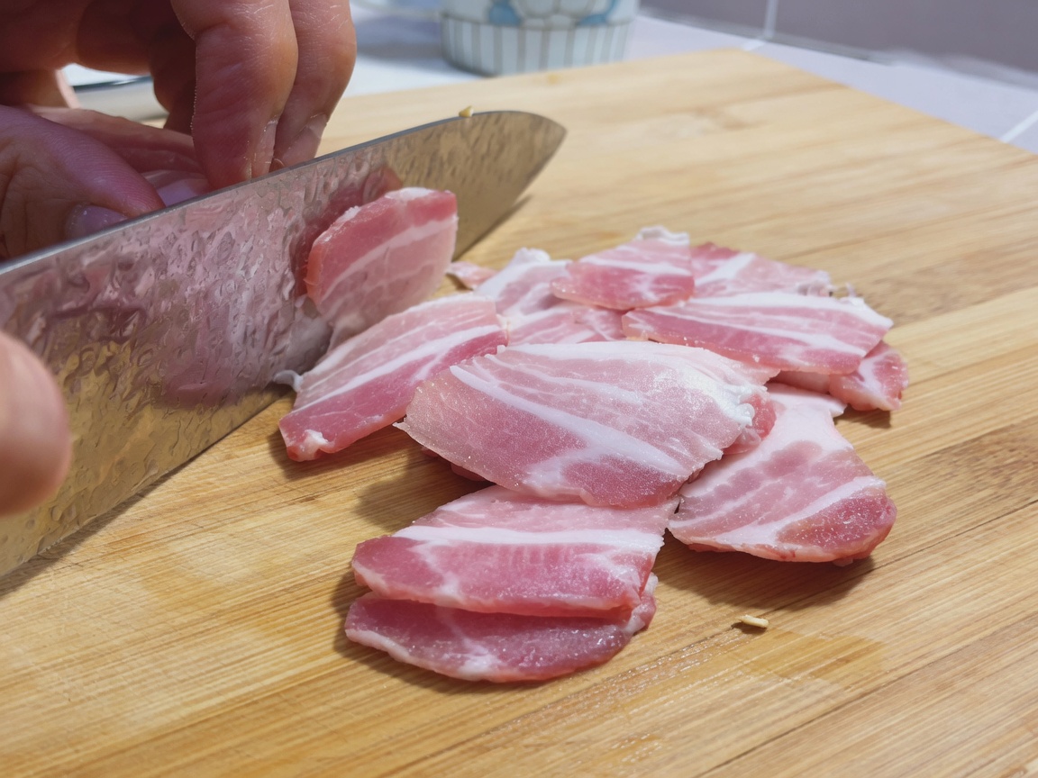 Thin slices of raw pork belly arranged on a wooden cutting board next to a large chefs knife.