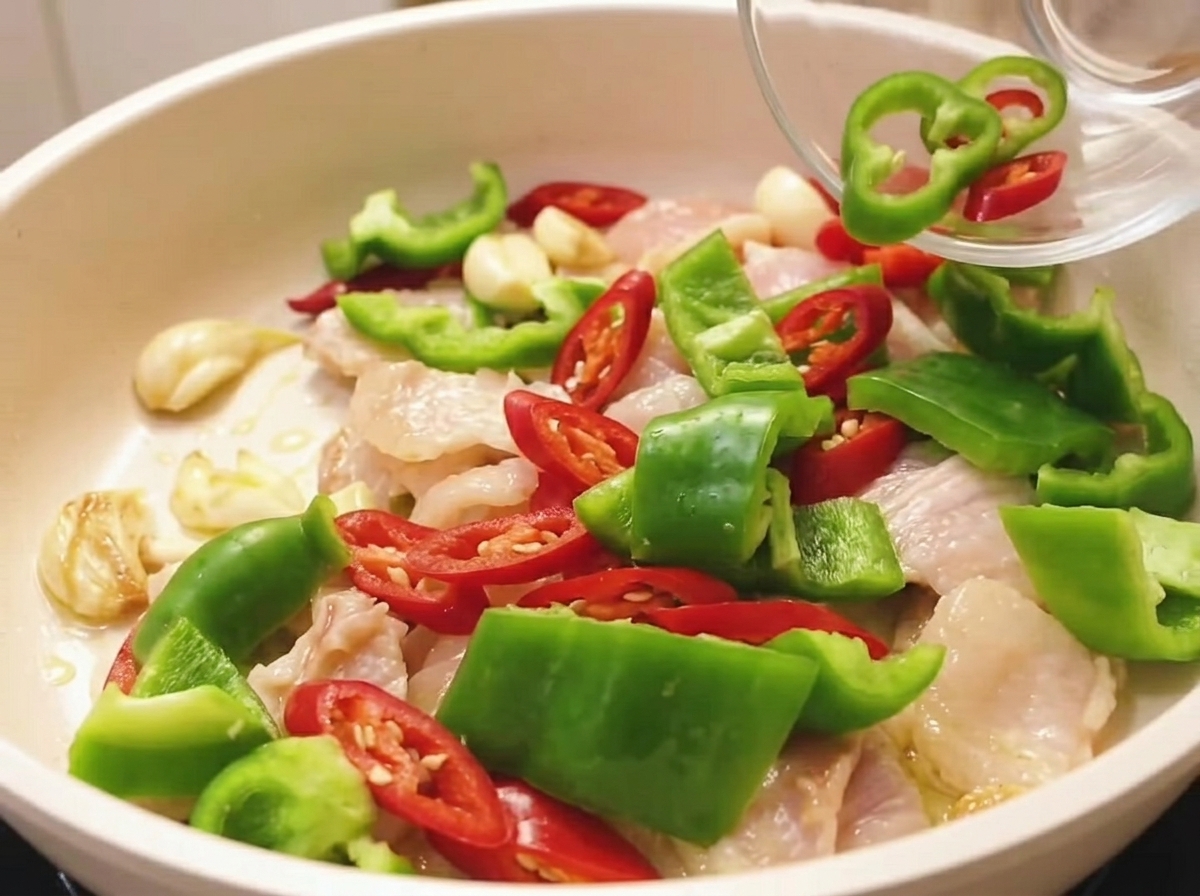 Freshly chopped red and green bell peppers being poured from a small glass bowl into a pan of partially cooked sliced pork.