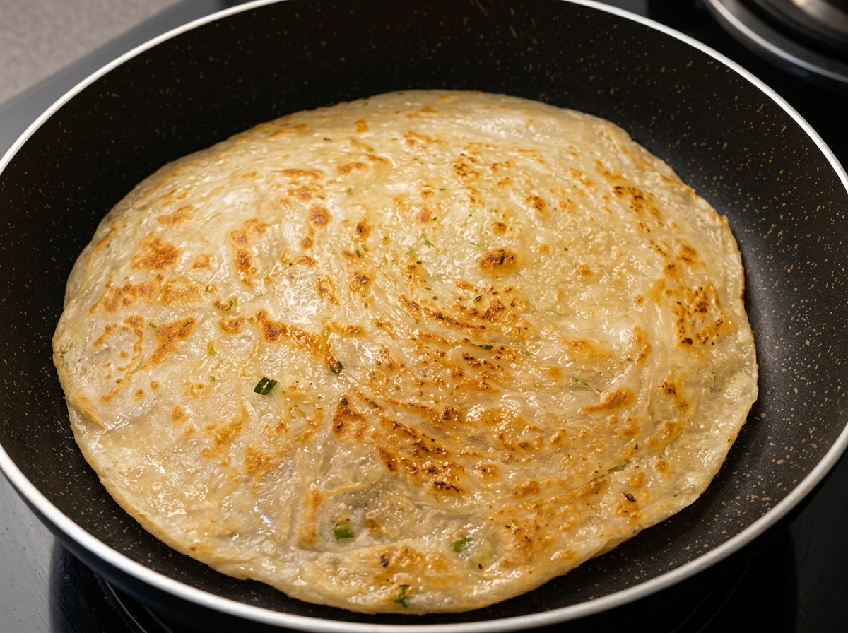 A round scallion flatbread frying in a dark non-stick pan, showing a golden, textured surface.