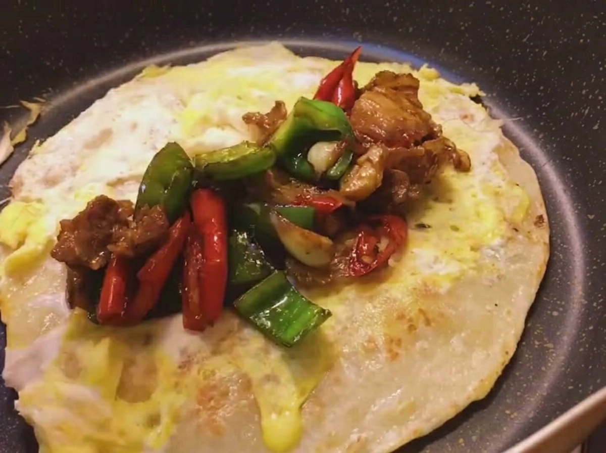 Spicy stir-fried pork and pepper filling being placed onto a golden-brown scallion flatbread with an egg cooked on top.